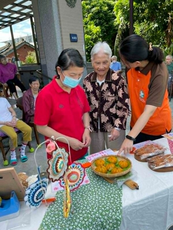 小太陽市集裡，工作人員正和跟長者介紹說明市集販售的南瓜包，南瓜包的餡料是長者一起參與製作而成。（圖片來源/小太陽）