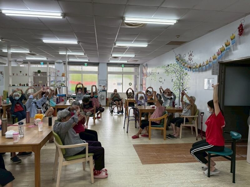 松柏樂復易的日照中心內，照服員正帶領長者進行坐站運動。（圖片來源／松柏樂復易）